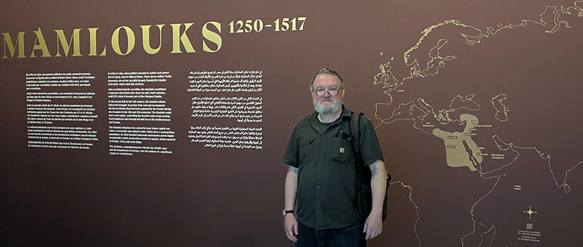 A man with a gray beard, glasses, and casual clothing stands in front of a large exhibition wall titled "Mamlouks 1250–1517." The wall features Arabic calligraphy above the title, explanatory text in English and Arabic, and a gold world map highlighting the Mamluk Empire region in the Middle East and North Africa.