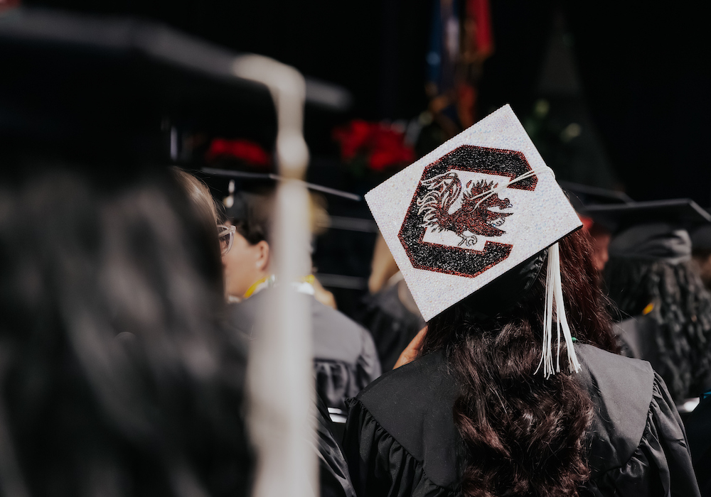 person in graduation robes wearing a bedazzled graduation cap with a gamecock and a block C on it