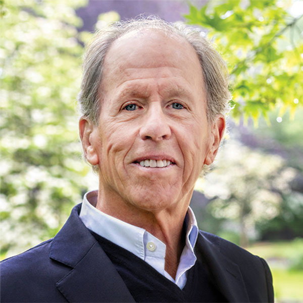 Portrait of Peter McCausland outdoors, wearing a dark jacket over a collared shirt and sweater, smiling slightly with soft natural light and green foliage in the background. 